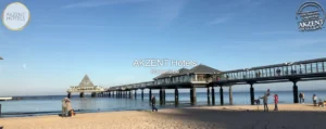 A scenic view of a beach with a pier in the distance, featuring people enjoying the day, under clear blue skies, with the AKZENT Hotels logo overlayed on the image.